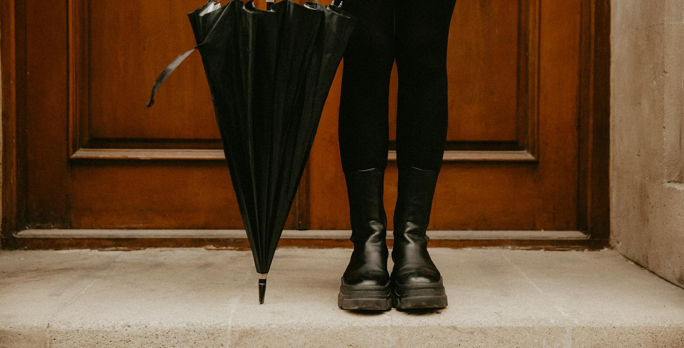 A person wearing black pants and chunky black shoes stands in front of a wooden door, holding a black umbrella